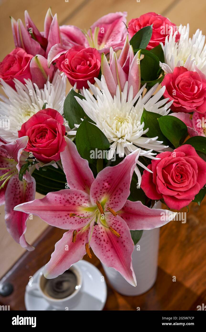 Une photographie recadrée de vase de fleurs roses et blanches dans une cruche en métal rose sur une petite table en acajou avec une tasse de café Banque D'Images