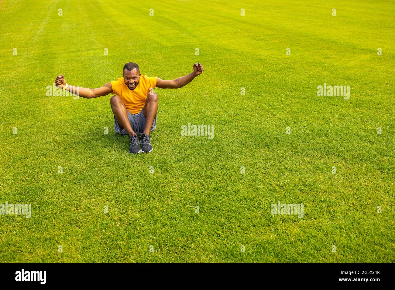 Sorti jeune athlète afro-américain dans des vêtements de sport jaune vif, assis sur l'herbe, souriant célébrant le succès, la victoire. Un mode de vie sain Banque D'Images