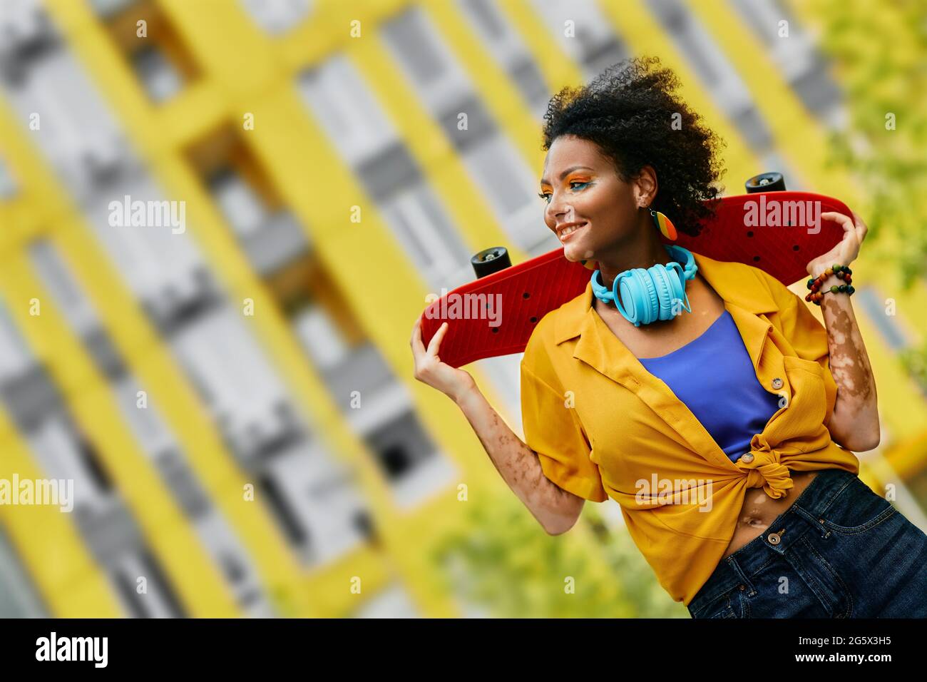 Belle femme aux cheveux bouclés tient le skateboard entre les mains tout en marchant dans une ville moderne en profitant des loisirs d'activité estivale Banque D'Images
