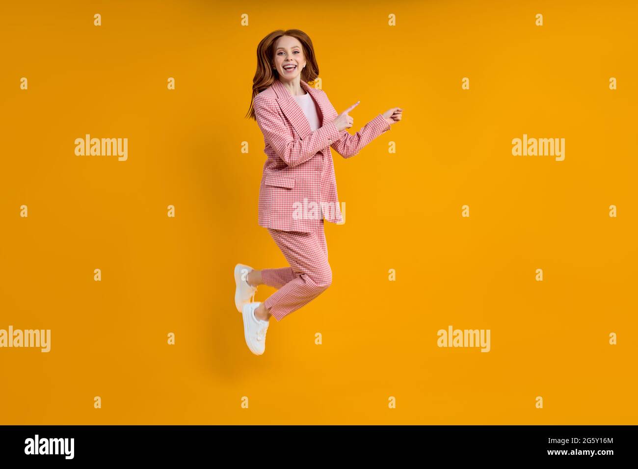 Femme d'affaires réussie en costume rose sautant doigt d'index sur le côté, isolé sur fond jaune, shopping. Portrait. Espace de copie pour publicité Banque D'Images