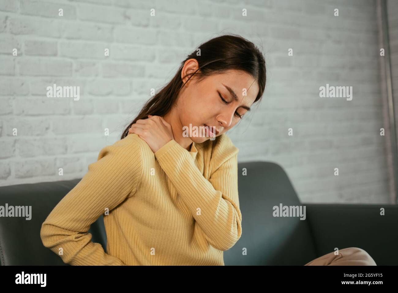 Jeune femme asiatique souffrant de douleurs à l'épaule tout en étant assise et en se reposant sur un canapé à la maison dans des vêtements décontractés avec les yeux fermés Banque D'Images