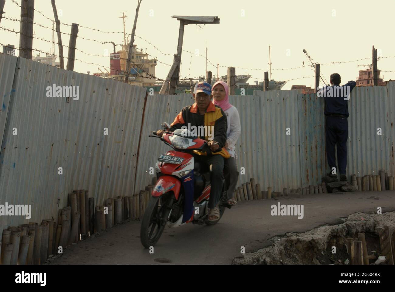Les automobilistes qui conduisent sur une route étroite sur le côté de la clôture d'un chantier naval, près du village côtier de Marunda à Cilincing, Jakarta, Indonésie. Banque D'Images
