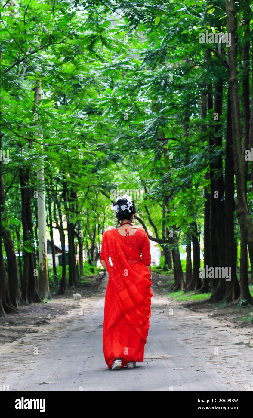 Jeune femme portant une saree rouge marchant sur une route verte en asphalte. La fille marche le long du chemin dans les bois à la lumière. Banque D'Images