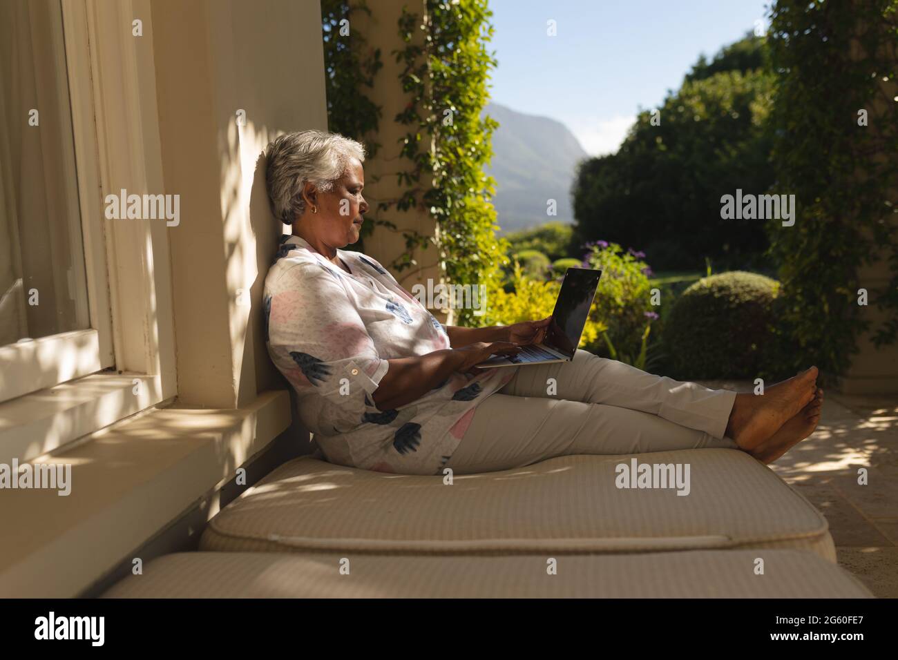 Femme afro-américaine senior utilisant un ordinateur portable sur une terrasse ombragée dans un jardin ensoleillé Banque D'Images