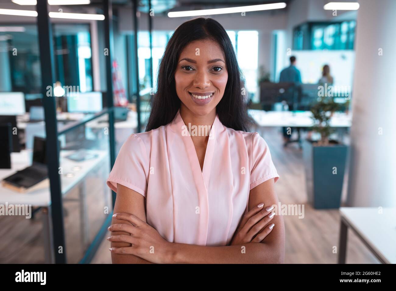 Portrait d'une femme d'affaires de race mixte souriant avec les bras croisés avec des collègues en arrière-plan Banque D'Images
