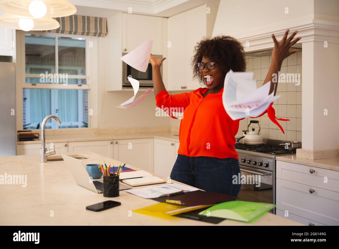 Bonne femme afro-américaine travaillant dans la cuisine avec un ordinateur portable, célébrant le succès, jetant des papiers Banque D'Images