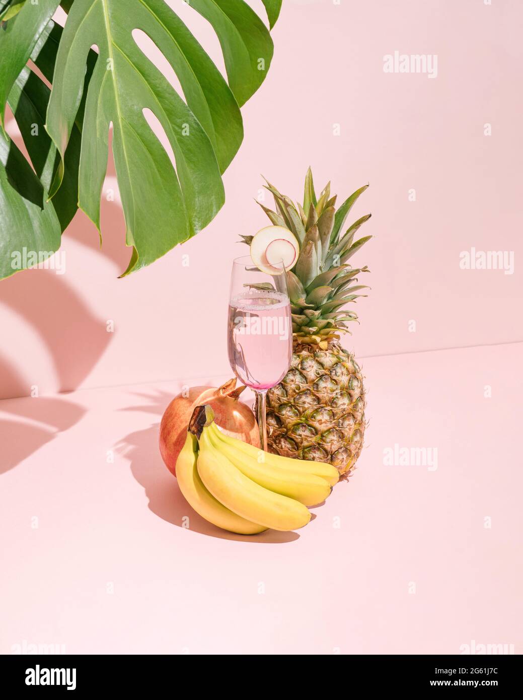 Bananes fraîches, ananas, grenade et jus de rafraîchissement en verre sur fond rose pastel. Ombre végétale verte naturelle. Scène tropicale d'été. Banque D'Images