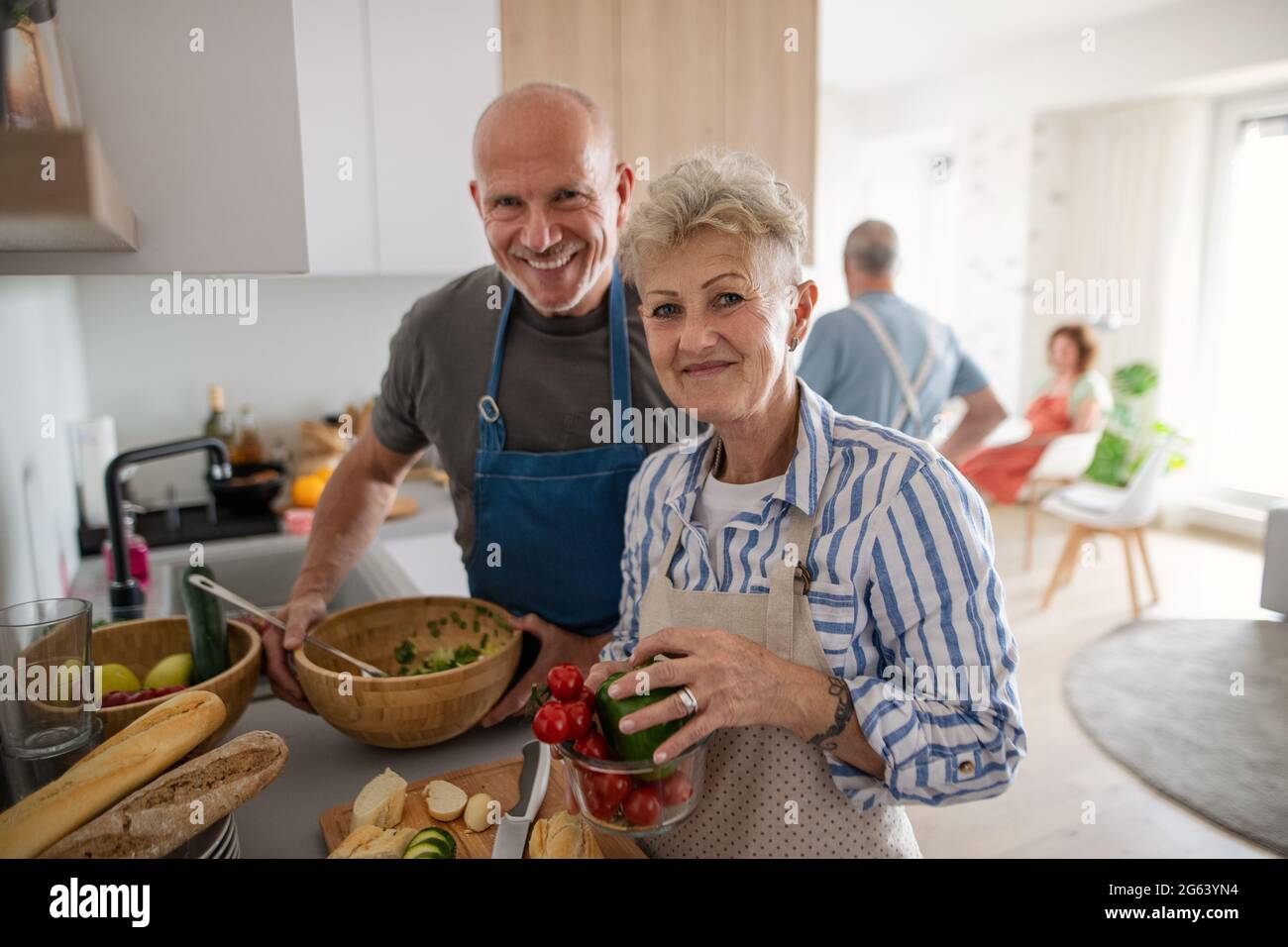 Couple senior avec des amis qui ont la fête à l'intérieur, regardant l'appareil photo lors de la cuisine. Banque D'Images