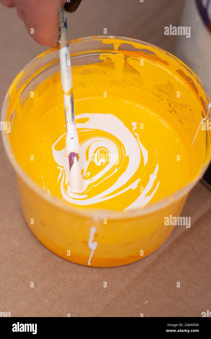 macro main mélange la peinture jaune et blanche avec un bâton de bois dans un récipient en plastique Banque D'Images