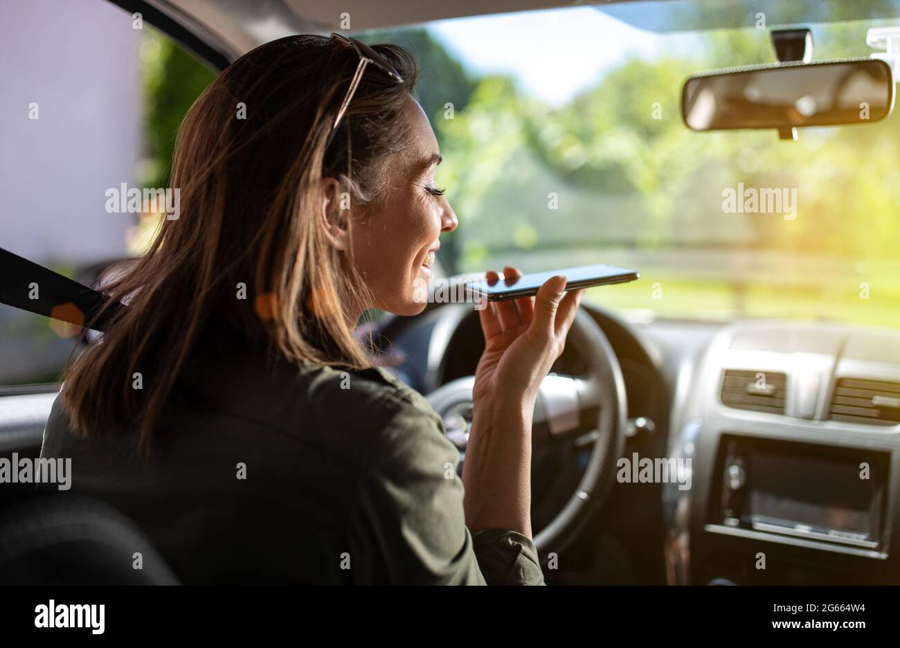 Jeune femme assise sur le siège du conducteur et portant sa ceinture de sécurité par beau temps. Jolie fille utilisant un téléphone intelligent sur le haut-parleur souriant. Banque D'Images