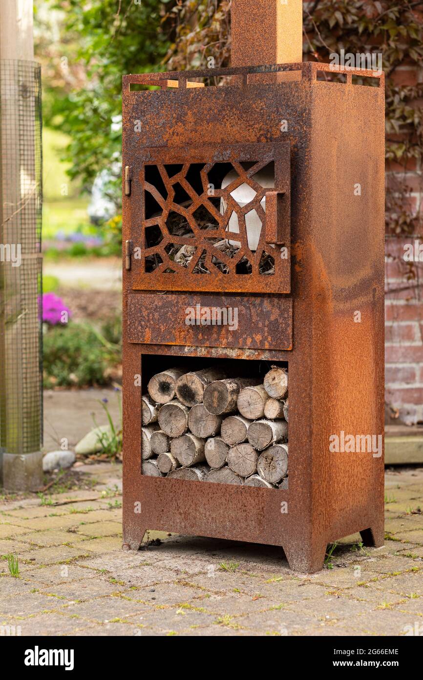 Vieux poêle à bois rouillé avec bois de bois debout dans un beau jardin idyllique avec des tondeuses, des arbres et de la verdure pendant une journée ensoleillée créant un paysage conscient Banque D'Images