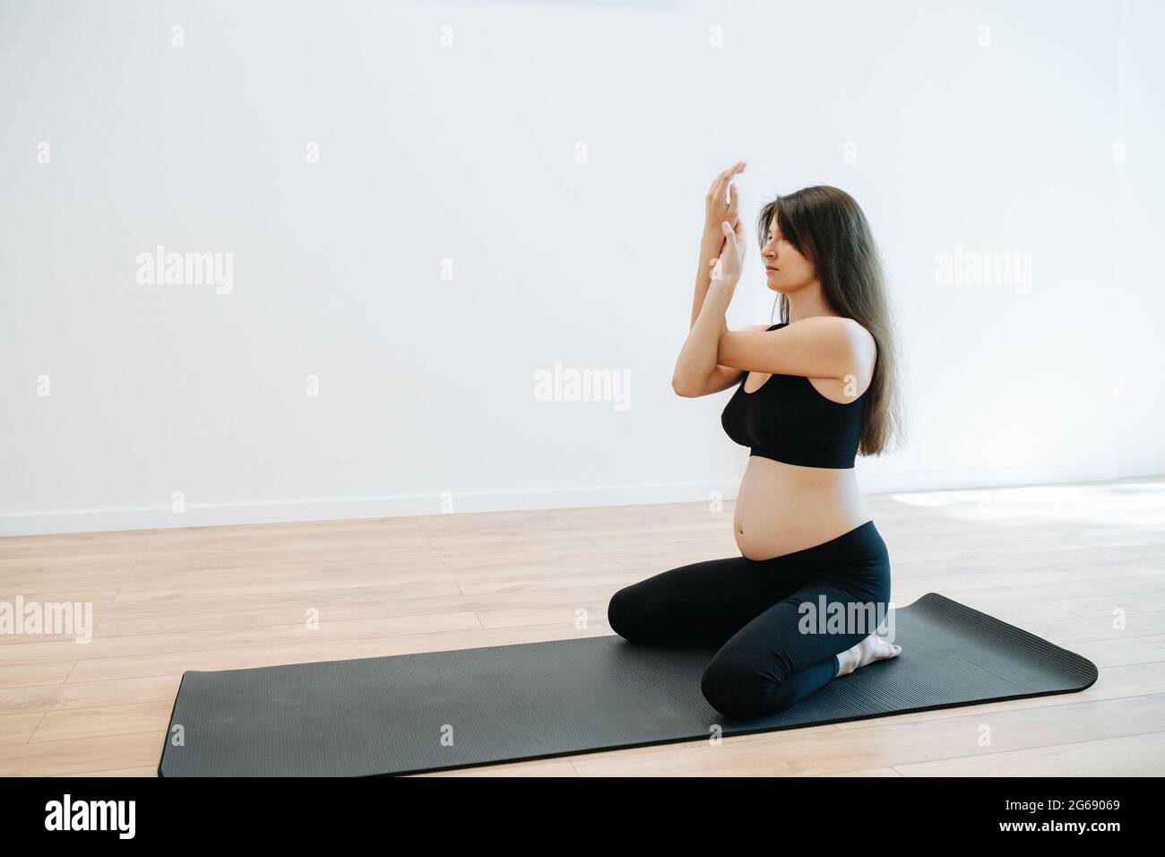 Femme enceinte aieuse assise sur un tapis à genoux avec un dos droit, pratiquant le yoga seul dans un grand studio. Ses bras s'emmêlent. Elle porte du noir Banque D'Images