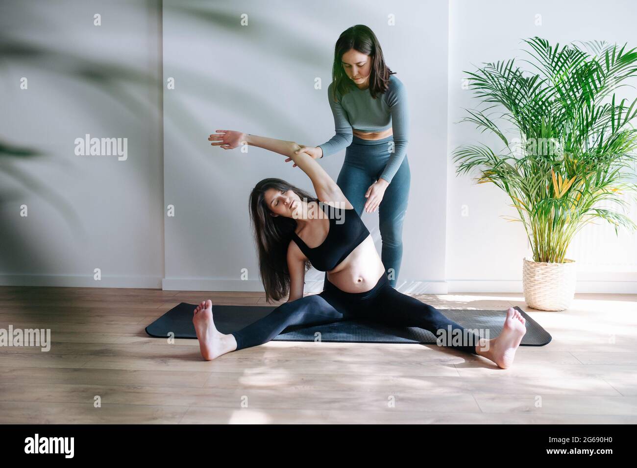 Coach aidant la femme enceinte à pratiquer le yoga, ils font des étirements latéraux. La femme est assise sur un tapis avec des jambes étalées dans un grand studio. Elle porte bl Banque D'Images