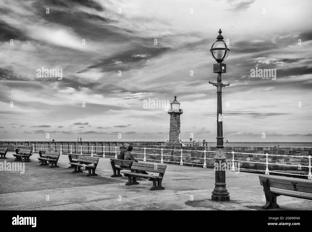 Un couple s'assoit sur un banc avec un lampadaire orné d'un côté. Un quai avec un phare est à mi-distance et un ciel avec nuage est au-dessus. Banque D'Images