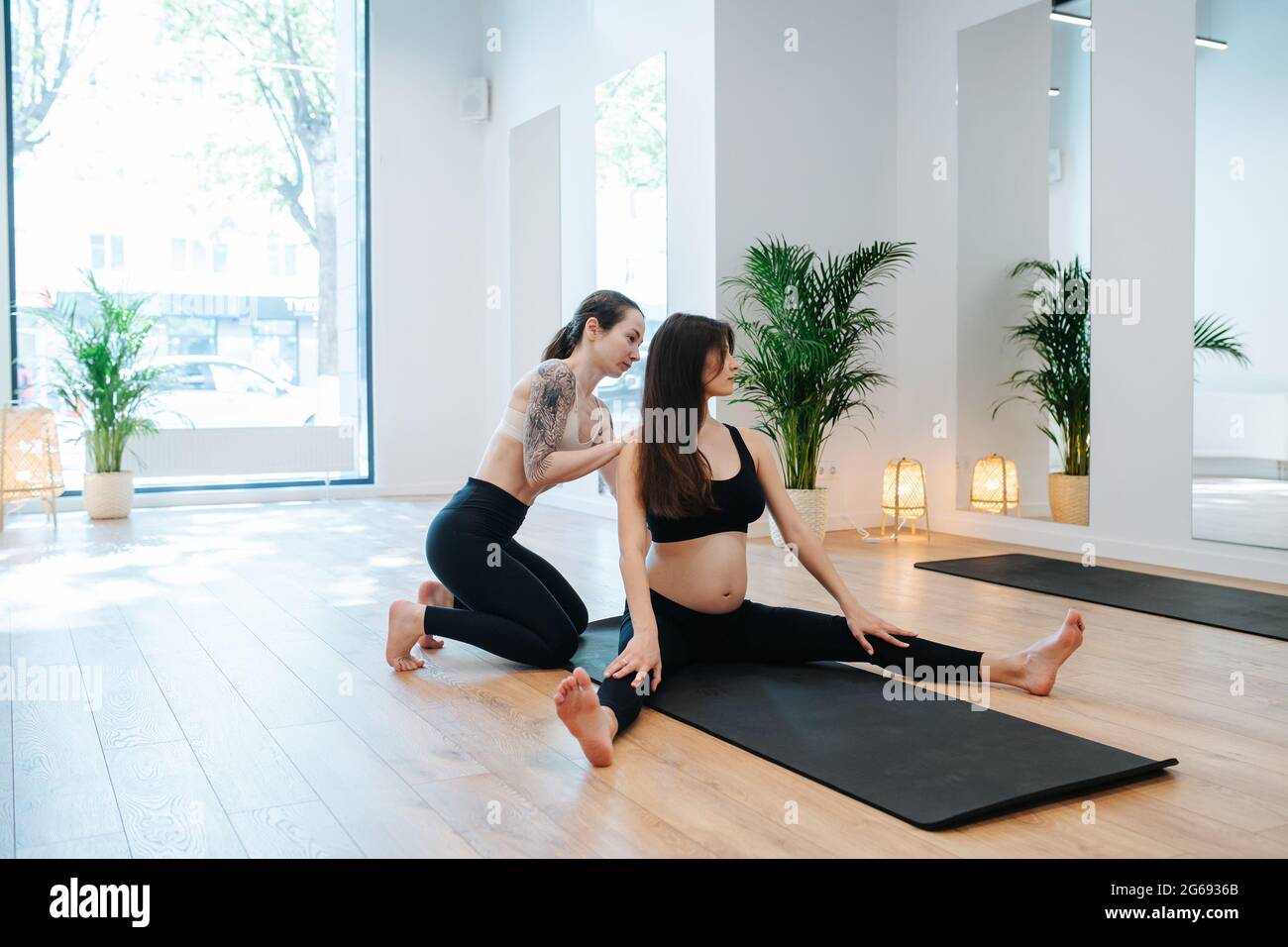 Instructeur aidant la femme enceinte à pratiquer le yoga, redressant son dos. La femme est assise sur un tapis avec des jambes étalées dans un grand studio. Elle porte Banque D'Images