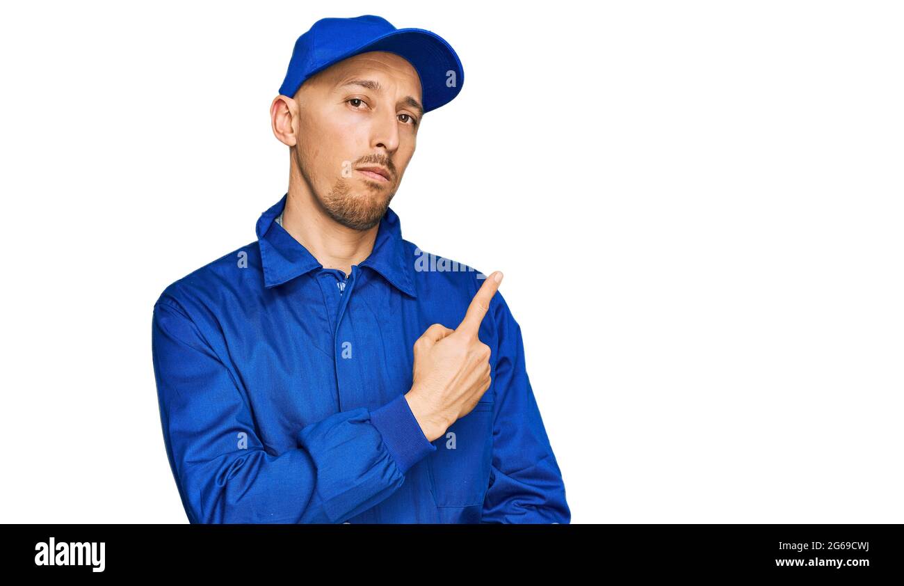 Homme chauve avec la barbe portant la combinaison de construction uniforme pointant avec le doigt de main sur le côté montrant la publicité, sérieux et calme visage Banque D'Images