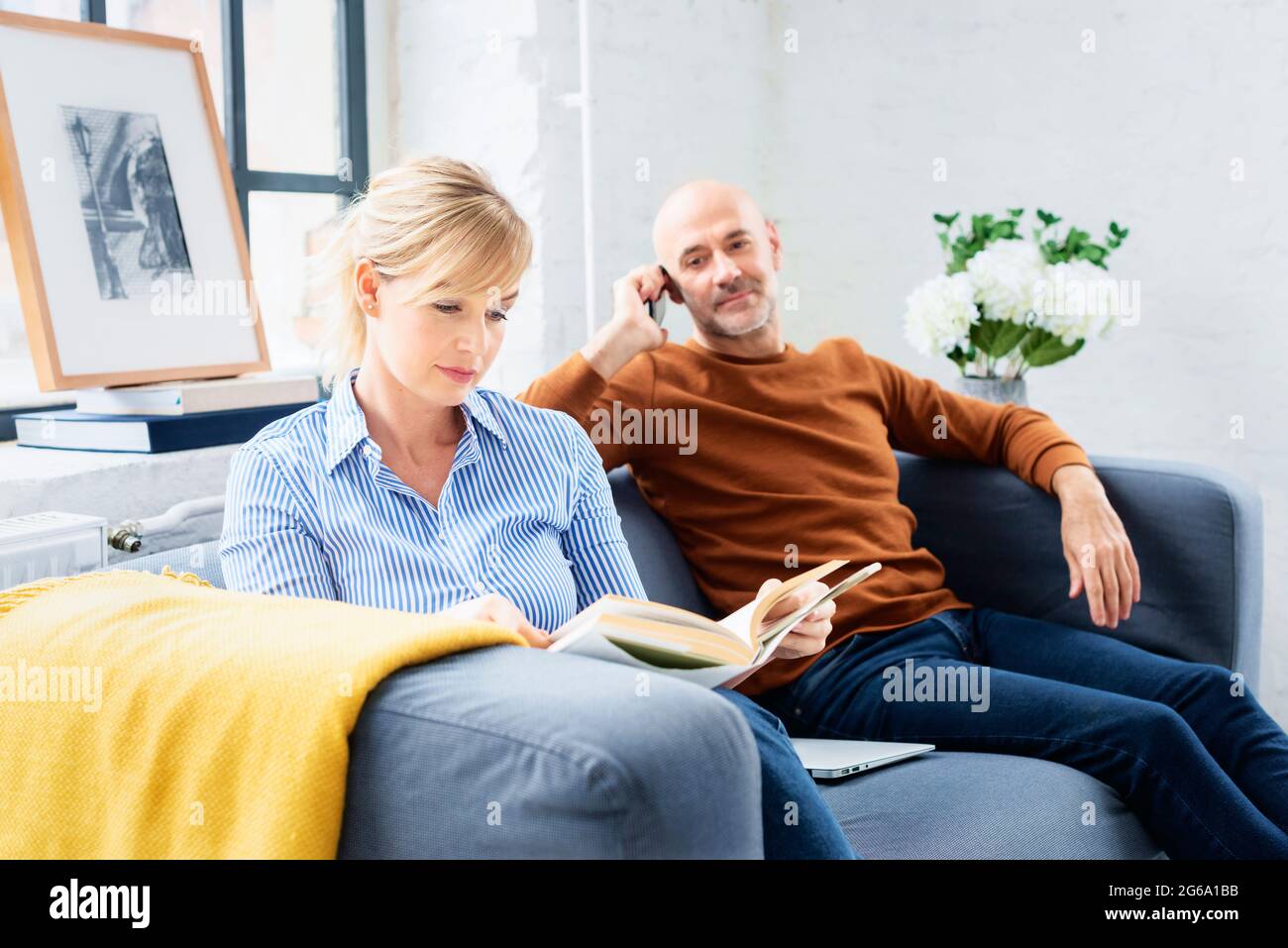 Belle femme lisant un livre tandis que son mari assis à côté de lui sur le canapé et parlant avec quelqu'un sur son téléphone mobile. Banque D'Images