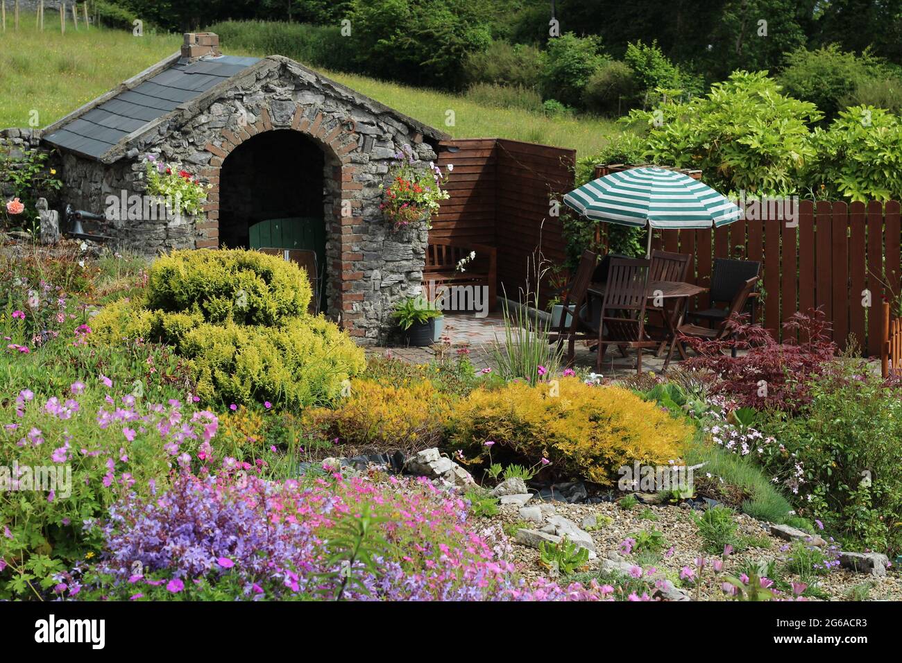 Jardin en Irlande en été avec rocaillerie en premier plan avec fleurs en fleur, petit bâtiment en pierre forge et coin salon avec patio Banque D'Images