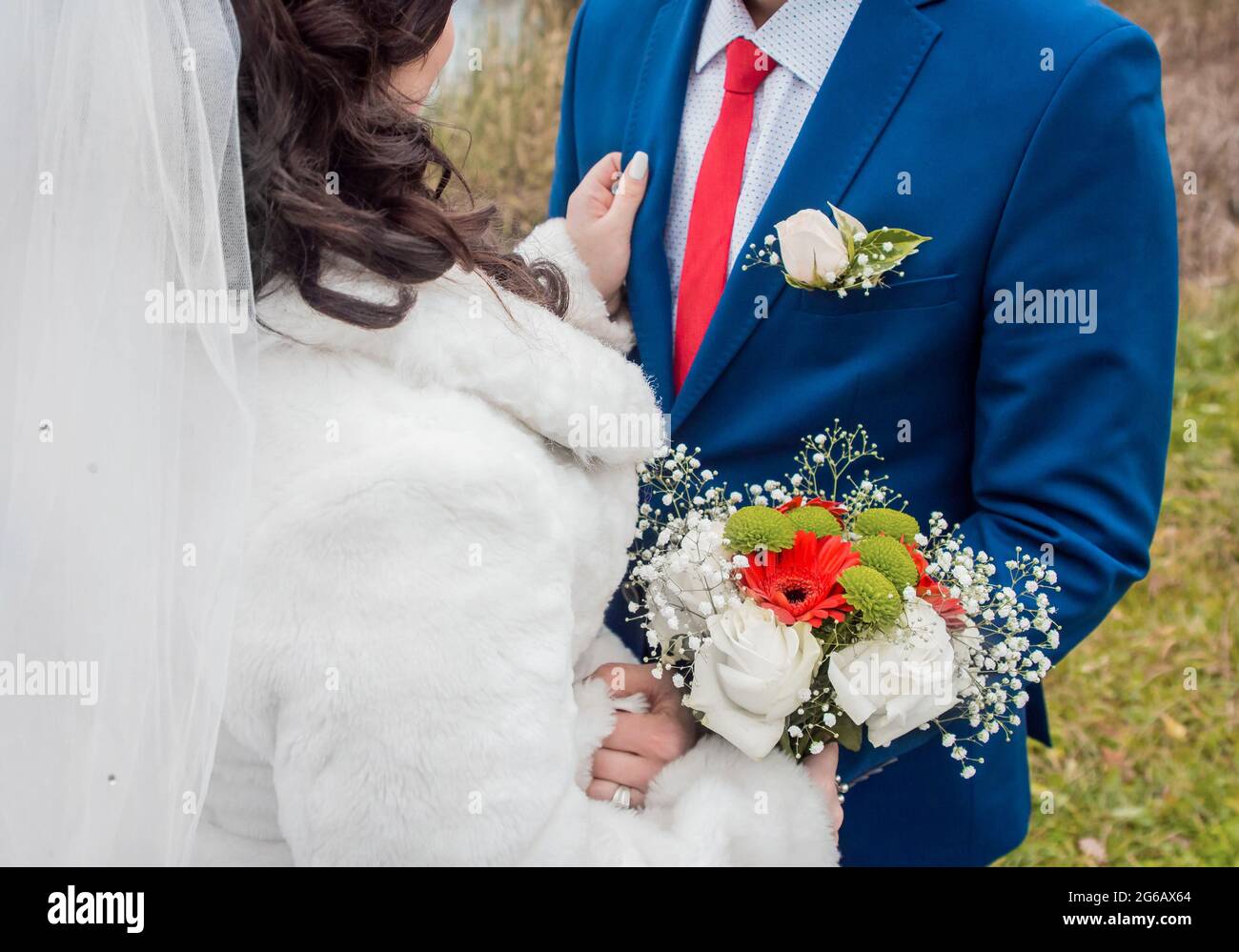 La mariée dans un voile dans un manteau blanc se tient en face du marié  dans un costume bleu, cravate rouge et chemise blanche avec un bouquet de  mariage de fleurs. La