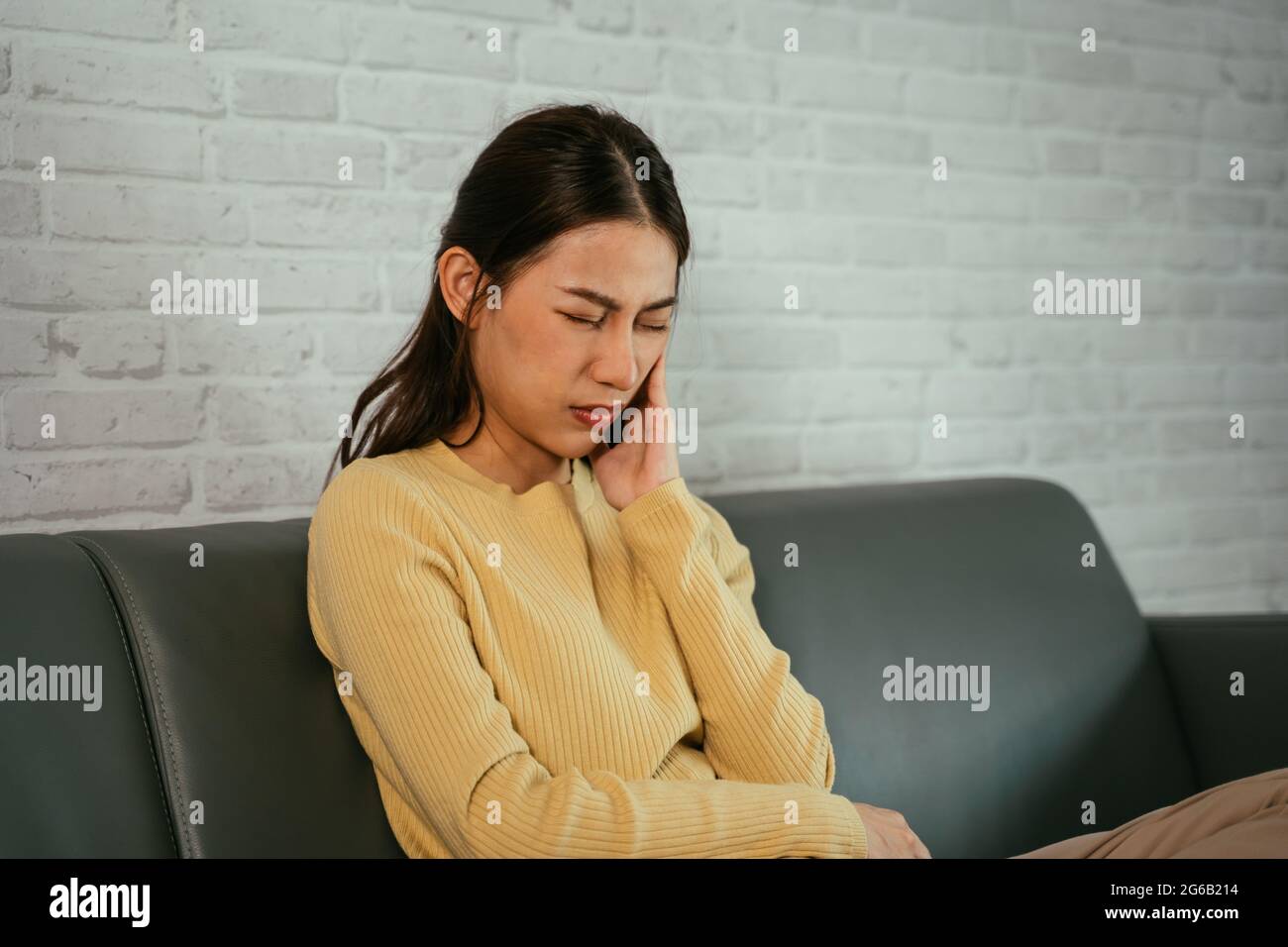 Femme asiatique assise sur un canapé avec les jambes pliées et les yeux fermés appuyant sur la joue comme souffrant de douleur dentaire due à la carie et à la cavité dans les dents à la maison Banque D'Images