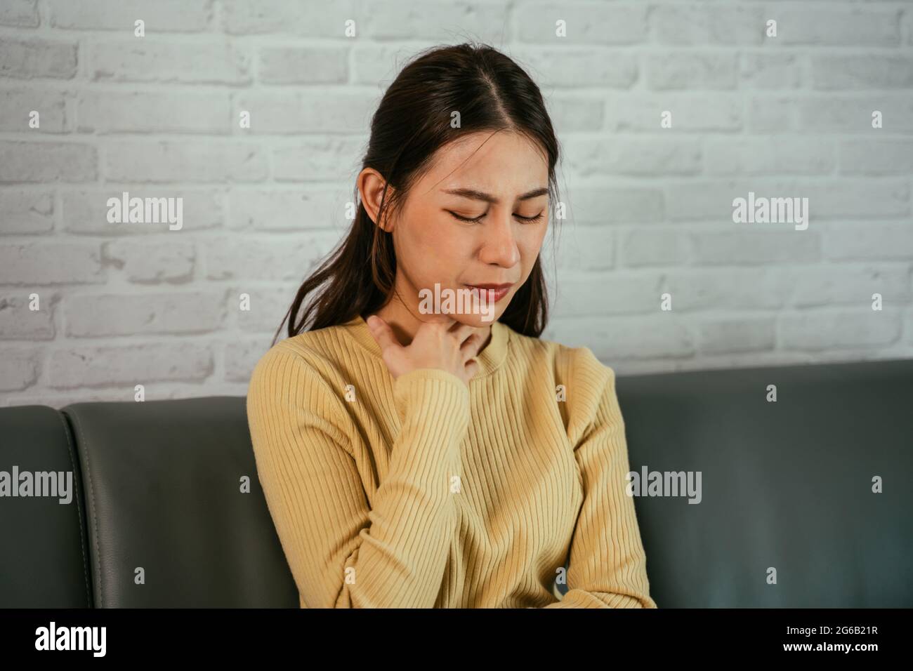 Jeune femme asiatique à la maison assise sur un canapé se reposant et touchant le cou en raison d'un mal de gorge avec les yeux fermés et un malaise Banque D'Images