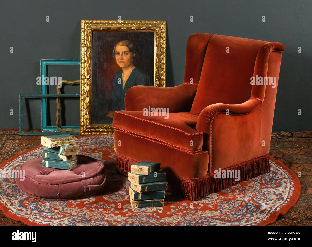 Fauteuil rouge vintage placé sur la moquette ornementale près de pouf et des piles de livres anciens contre des cadres avec portrait classique Banque D'Images