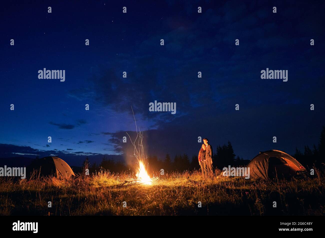 Magnifique paysage de nuit ciel nuageux sur une colline herbeuse avec tentes de camp, feu de camp et randonneurs. Les jeunes voyageurs, hommes et femmes, profitent de la vue sur le ciel bleu de nuit tout en se tenant près des tentes touristiques. Banque D'Images