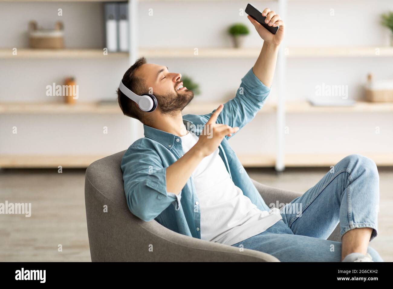 Jeune caucasien souriant dans un casque à l'écoute de musique ou de livre audio, assis dans un fauteuil à la maison Banque D'Images