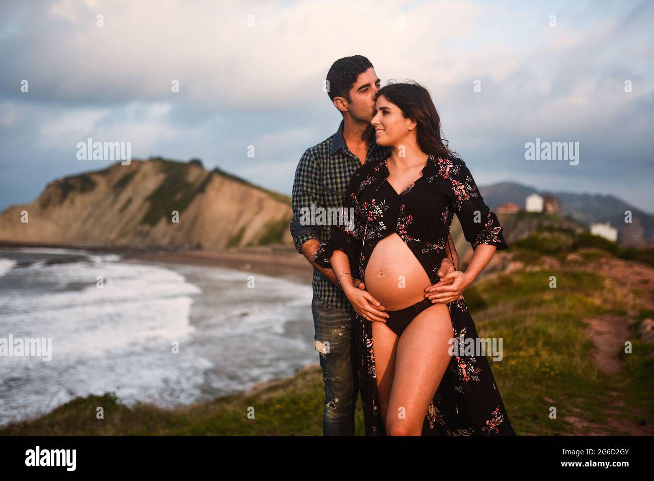 Homme aimant embrassant et embrassant une femme enceinte heureuse vêtue d'une tenue élégante révélant le ventre tout en se tenant debout sur la mer contre les montagnes Banque D'Images