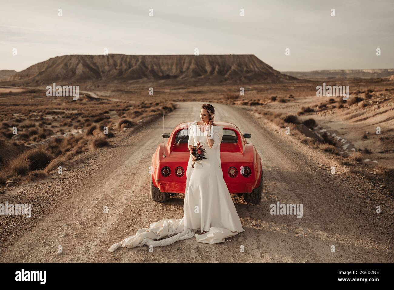 Une femme heureuse en robe blanche se tenant à l'écart près d'un véhicule rouge de luxe stationné sur une route poussiéreuse lors d'un voyage dans le parc naturel de Bardenas Reales à Nava Banque D'Images