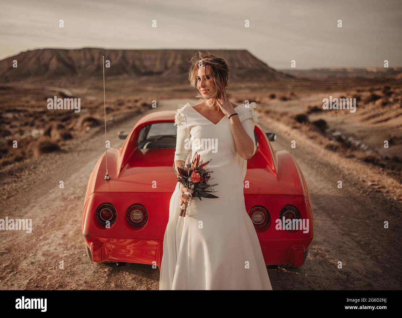 Une femme heureuse en robe blanche se tenant à l'écart près d'un véhicule rouge de luxe stationné sur une route poussiéreuse lors d'un voyage dans le parc naturel de Bardenas Reales à Nava Banque D'Images