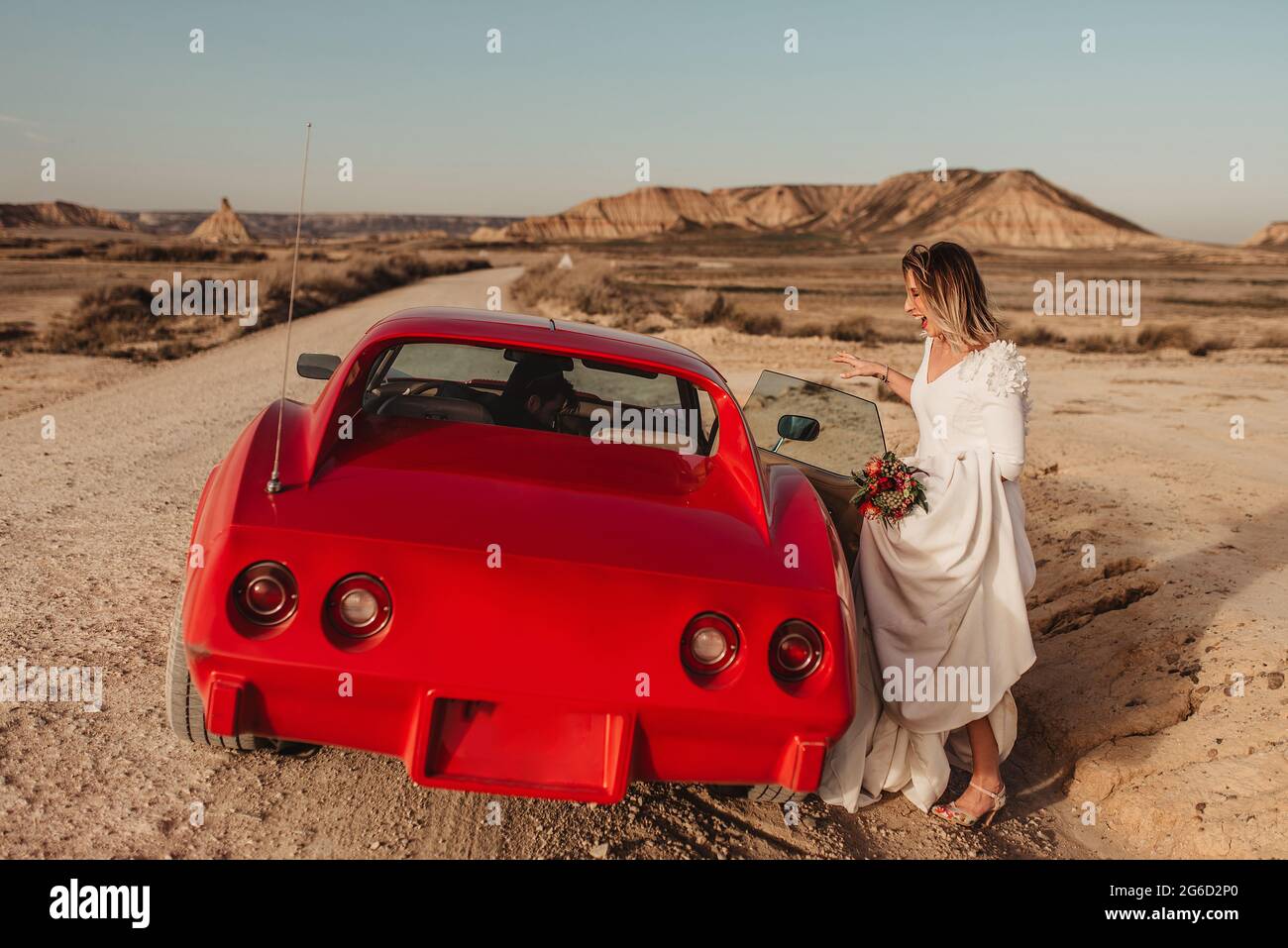 Femme pleine de corps heureuse en robe blanche se trouvant dans un véhicule rouge de luxe stationné sur une route poussiéreuse pendant le voyage dans le parc naturel de Bardenas Reales à Navarra, Banque D'Images