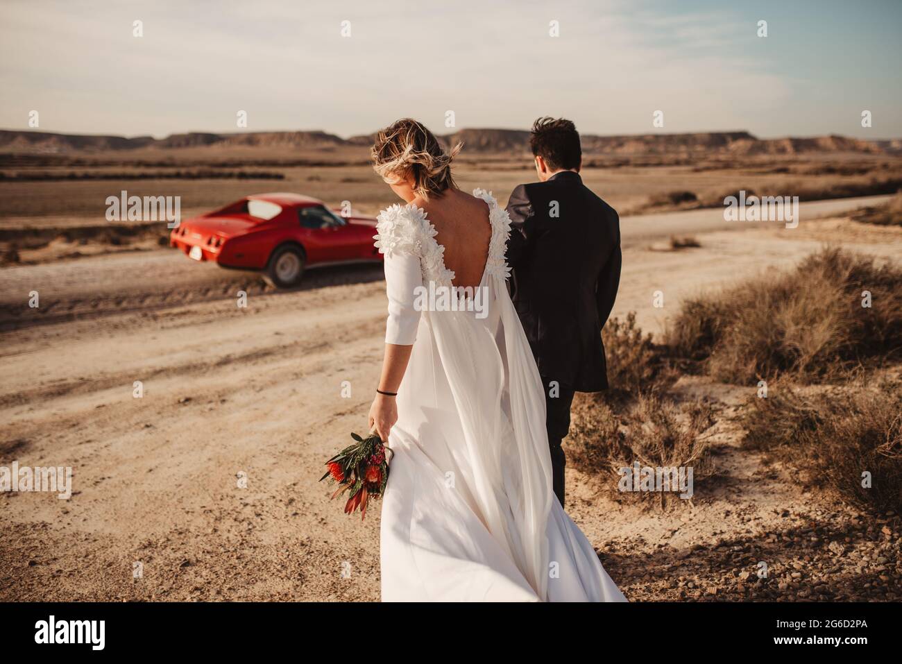 Vue arrière de la mariée et du marié anonymes se promenant ensemble vers une voiture rouge de luxe dans le désert du parc naturel de Bardenas Reales à Navarra, Espagne Banque D'Images