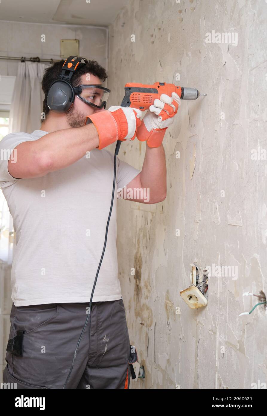 Constructeur forant des trous dans un mur avec une perceuse électrique sur le chantier de construction. Travailleur de la construction portant des gants de sécurité, des lunettes et des couvre-oreilles. Banque D'Images