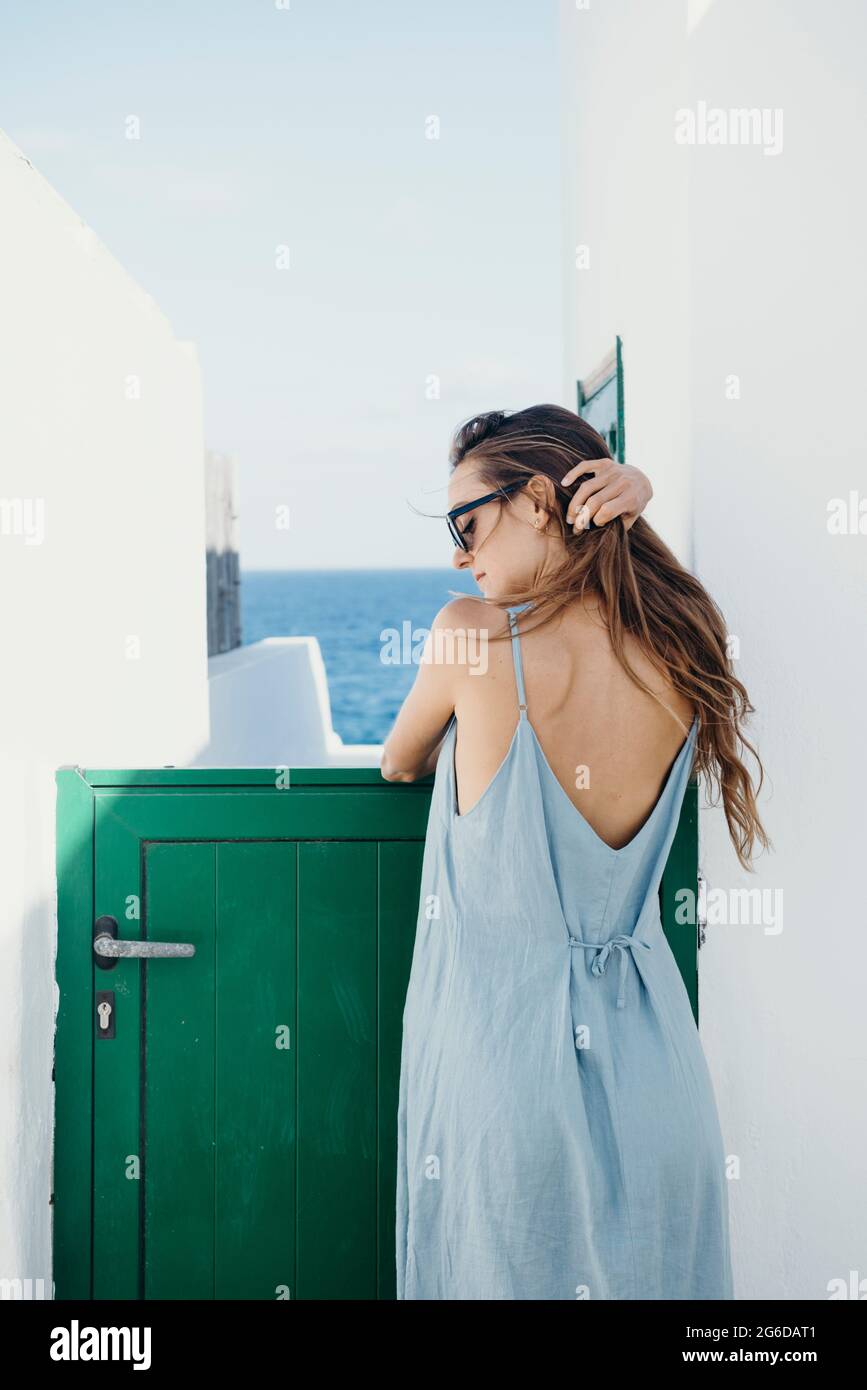 Vue latérale d'une jeune femme pieds nus dans une sundress et des lunettes  de soleil se penchant contre la porte verte d'une maison en regardant loin  tout en se relaxant seul en
