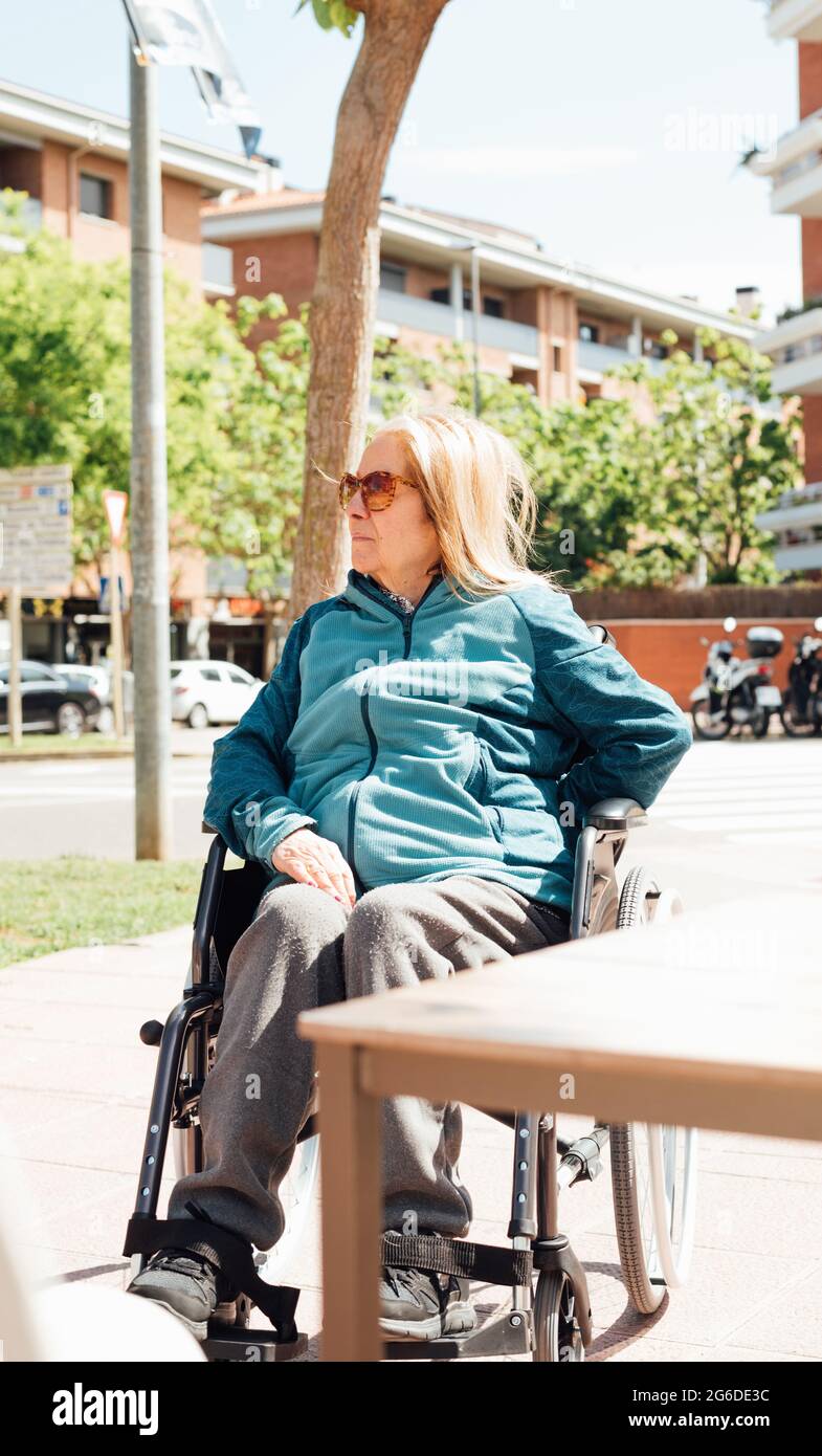 Une femme âgée pensive assise en fauteuil roulant dans la rue par beau temps et donnant sur l'extérieur Banque D'Images