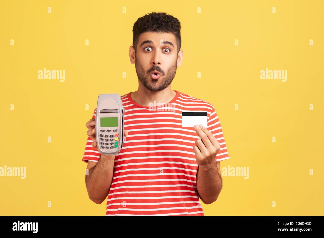 Homme barbu excité dans une tenue de t-shirt rayé montrant le terminal de paiement de pos et la carte de crédit, surpris par le cashback pendant les paiements sans espèces. Banque D'Images