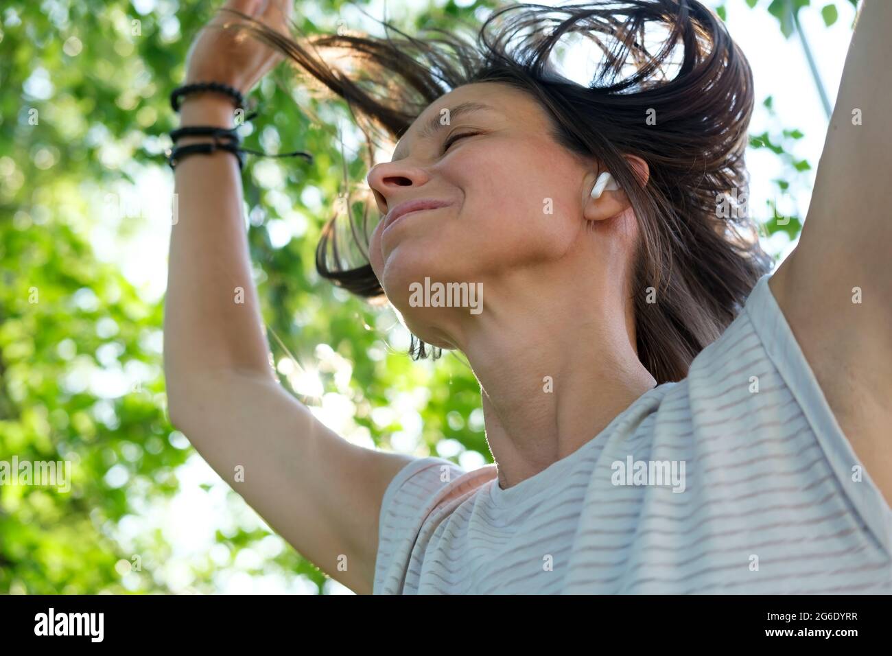 Une femme heureuse écoute de la musique dans des écouteurs sans fil et danse, avec ses cheveux en vol, un jour ensoleillé d'été. Banque D'Images
