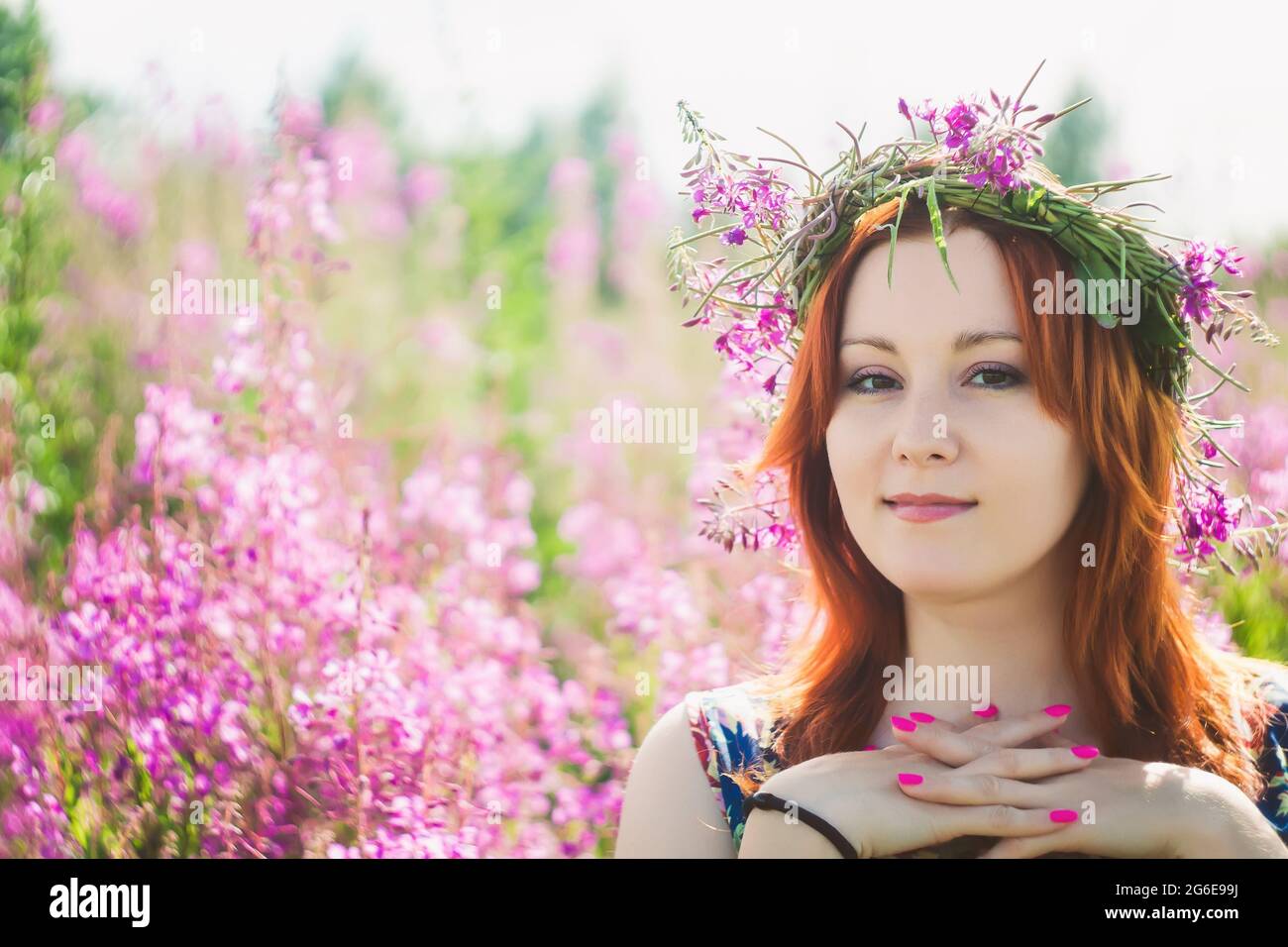 Jeune belle femme aux cheveux rouges avec une couronne de fleurs sauvages roses sur sa tête regarde l'appareil photo et sourit. Style été Banque D'Images