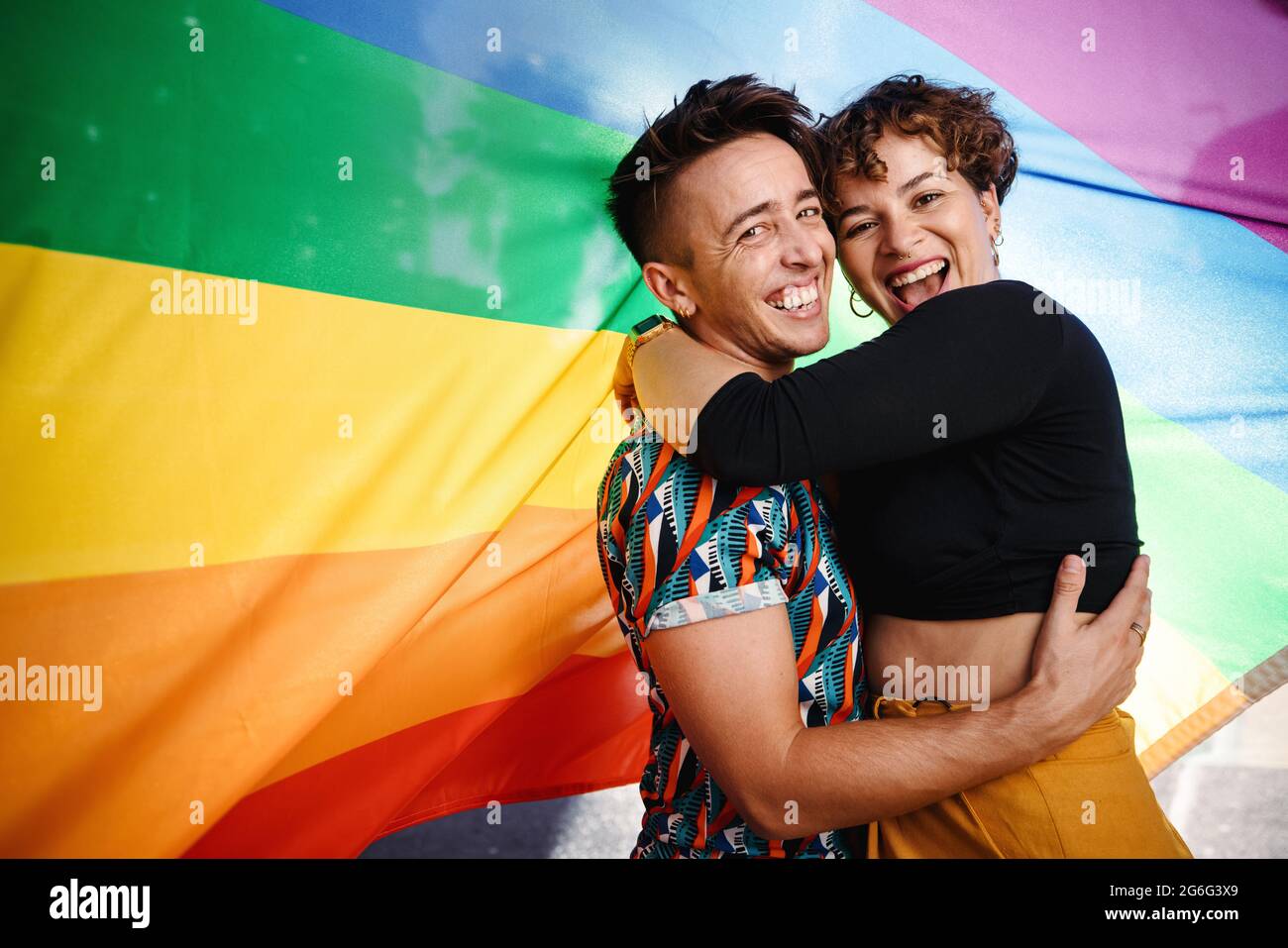 Couple queer souriant debout contre un drapeau de fierté arc-en-ciel. Un jeune couple LGBTQ s'embrasse les uns les autres tout en se tenant debout. Deux amoureux non-conformant Banque D'Images