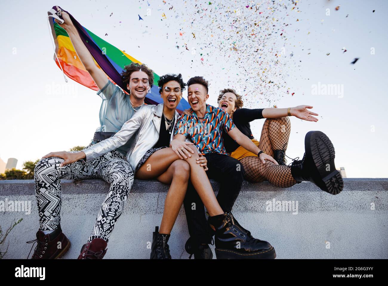Heureux reines célébrant la fierté gay. Groupe de quatre amis tenant le drapeau de fierté de l'arc-en-ciel tout en étant assis ensemble à l'extérieur. Membres joyeux de Th Banque D'Images