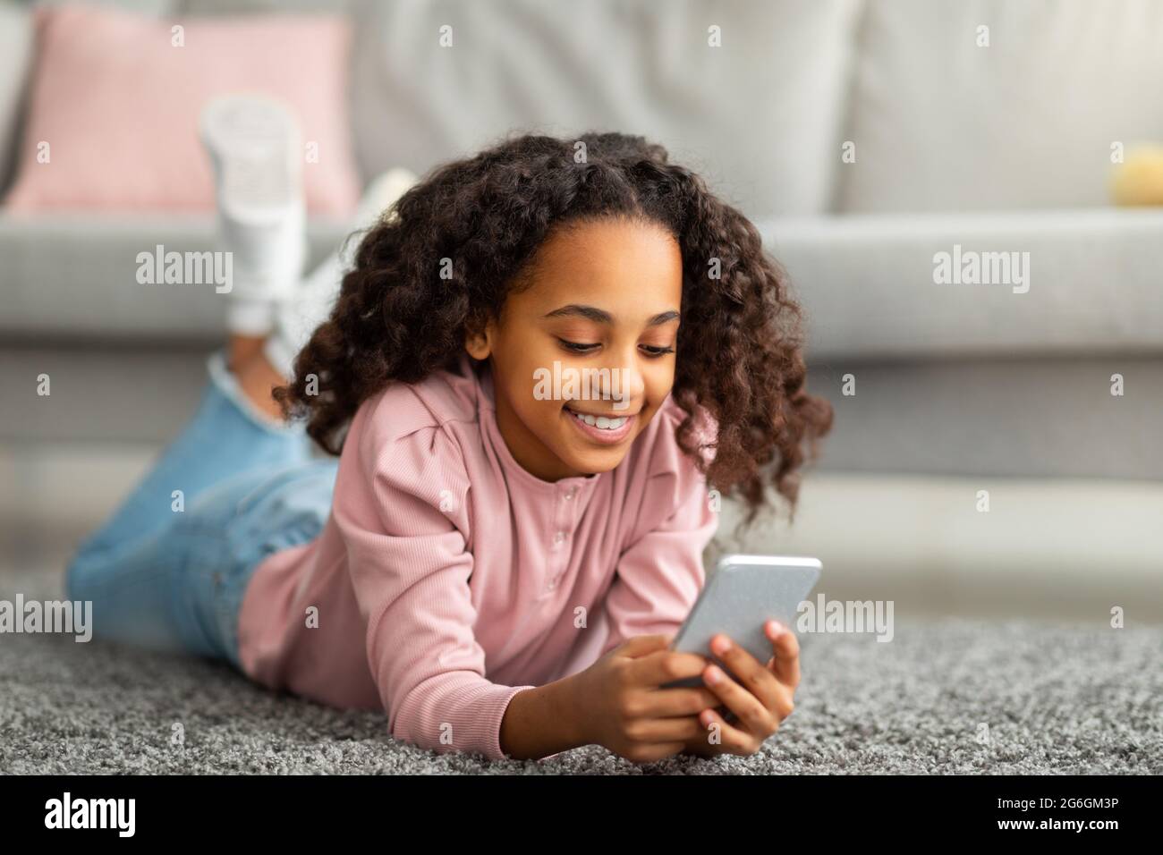 Natifs numériques. Une fille américaine africaine positive utilisant son smartphone, envoyant des SMS dans les médias sociaux, couchée sur le tapis de sol Banque D'Images