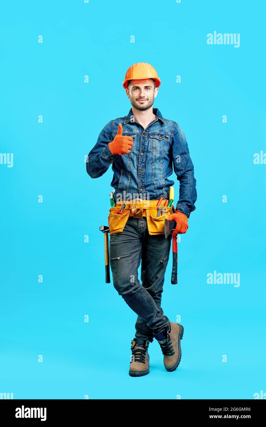 Portrait complet d'un homme de construction doué et confiant dans un casque de sécurité montrant le signe de pouce vers le haut sur fond bleu regardant la caméra Banque D'Images