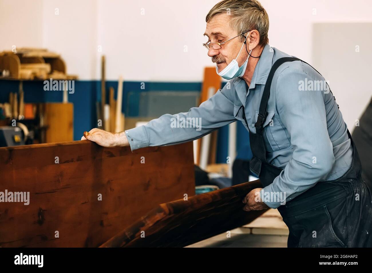 Un charpentier gris-cheveux avec des lunettes travaille avec du bois sur un établi dans une boutique de menuiserie. Un pensionné dans un masque médical au travail. Emploi et travail un homme âgé en vêtements de travail Banque D'Images