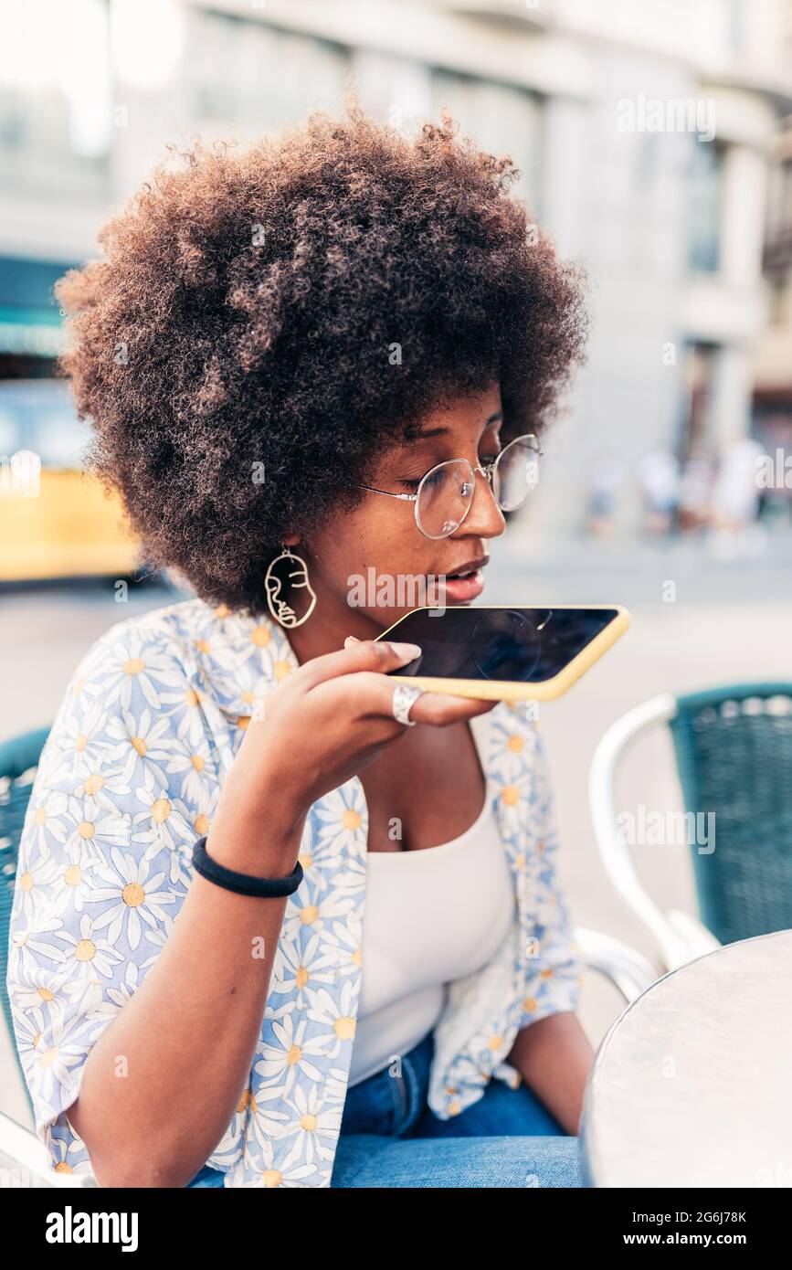 une jeune afro-américaine porte son téléphone près de sa bouche. Elle est dans une terrasse de bar. Concept de message vocal Banque D'Images