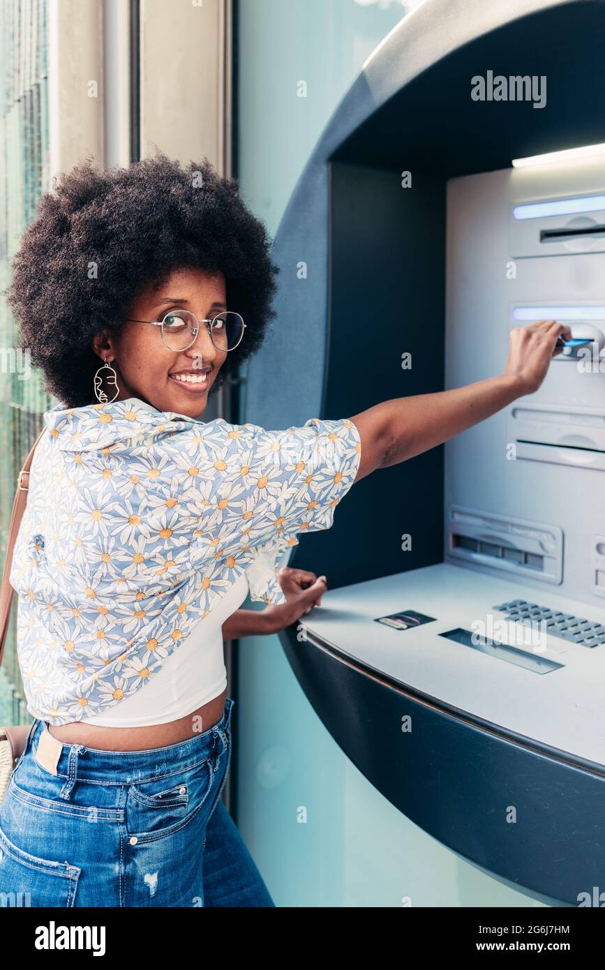 Portrait vertical d'une femme afro-américaine insérant une carte de crédit dans un guichet automatique. Elle regarde la caméra un sourire Banque D'Images