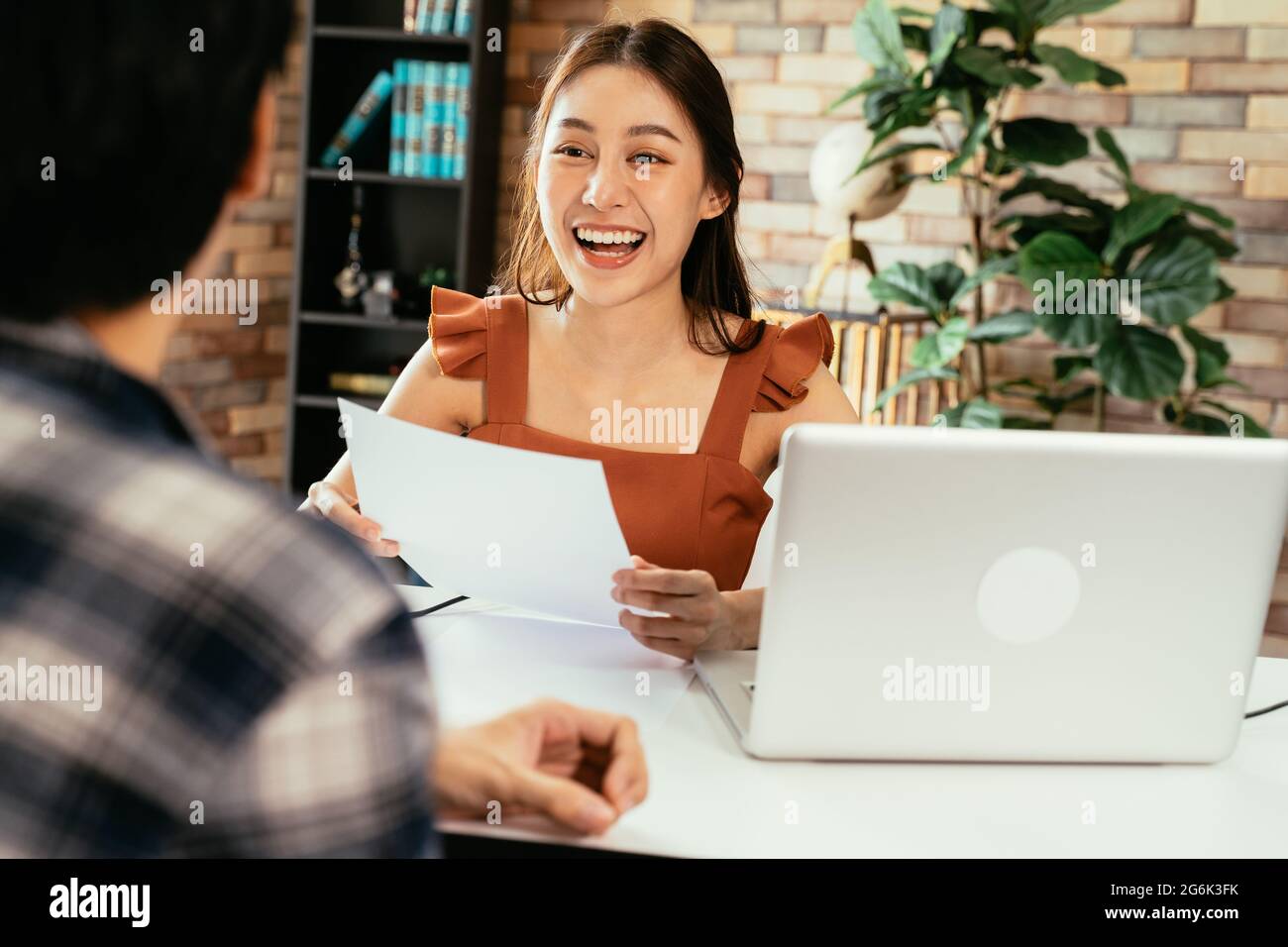 Une jeune femme asiatique joyeuse qui tient un questionnaire tout en interviewant un invité masculin dans l'espace de travail avec un micro et un ordinateur portable Banque D'Images