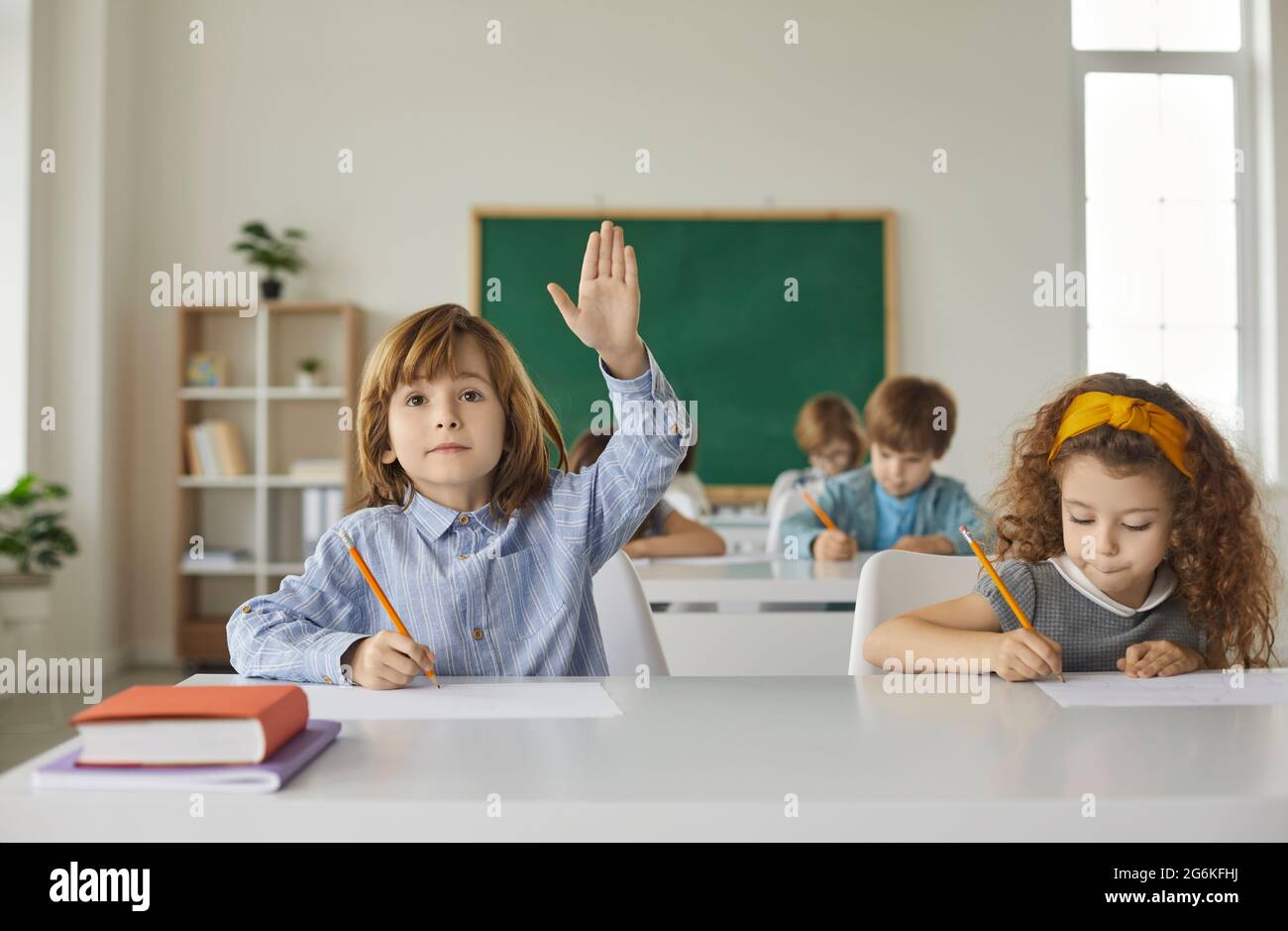Un garçon et une fille sont assis à un bureau d'école, écrivant, le garçon lève sa main vers le haut. Banque D'Images