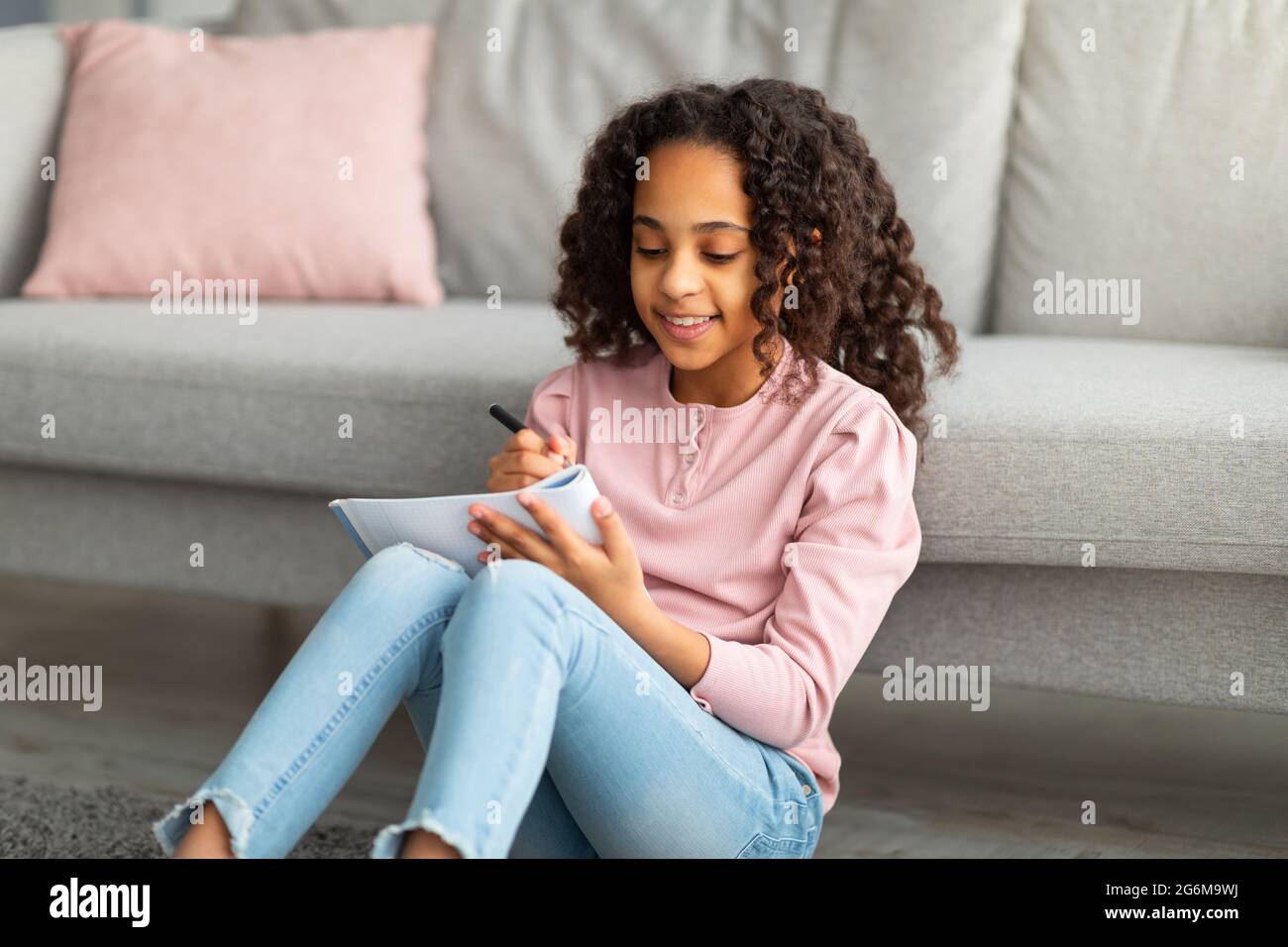 Gardez un journal personnel. Bonne jeune fille noire écrivant dans le carnet ses plans d'été et rêves, assis sur la moquette de plancher Banque D'Images