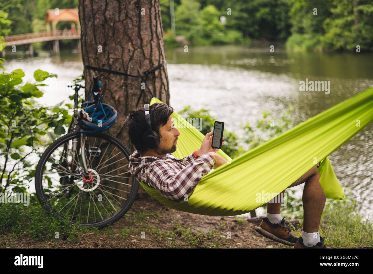 L'homme voyage à vélo, se détendre dans un hamac vert, surfer sur Internet sur un smartphone, écouter de la musique sur des écouteurs dans la forêt près du lac. Entrée cycliste Banque D'Images